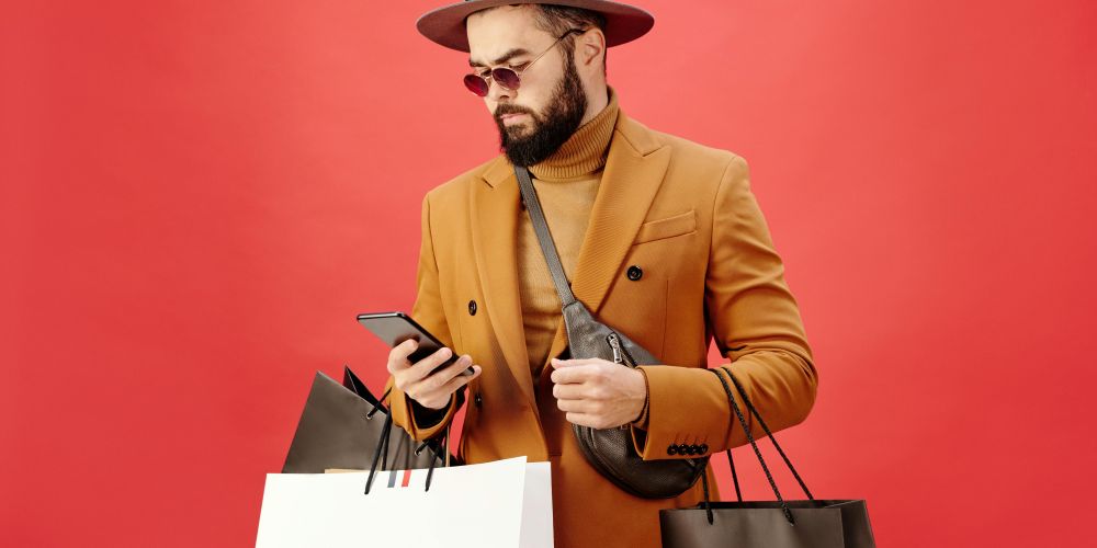 Foto von Max Fischer von Pexels, das eine Person mit Hut, Sonnenbrille, Pulli und Sakko zeigt, die Einkaufstüten trägt und auf ihr Smartphone schaut.