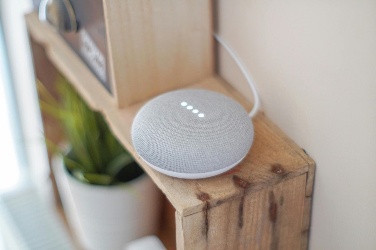 Ein grauer Google Home Lautsprecher auf einem Holzregal.