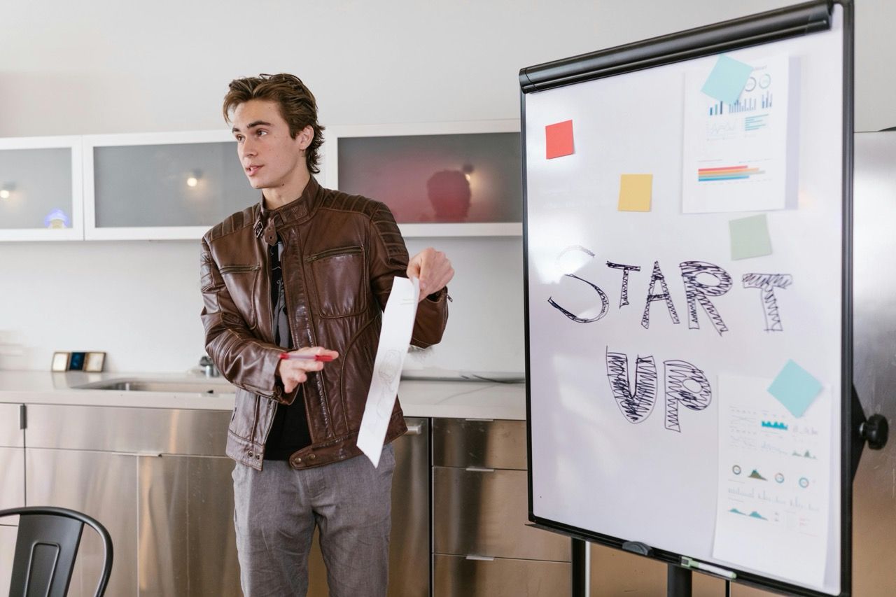 Junger Mann in Lederjacke präsentiert Startup-Idee an einem Whiteboard.